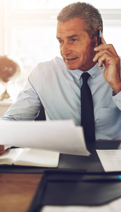 Smiling mature consultant discussing documents over the phone while working at his desk in an office